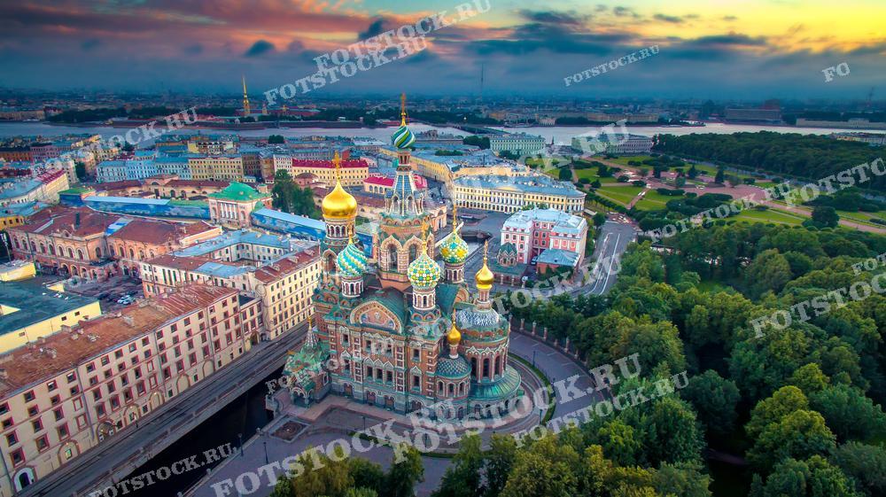 Фотография демонстрирует завораживающий вид на храм Спаса на Крови в Санкт-Петербурге, снятый с высоты в ранние утренние часы. Это изображение подойдет для использования в культурных журналах, туристических брошюрах и как визуальное пособие для обучения российской архитектуре и истории. Великолепные купола храма, окружённые зеленью парков и рекой Невой, создают неповторимый и яркий образ.